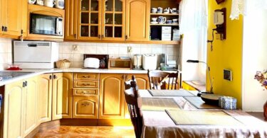 kuchyňská linka se spotřebiči, dřevěný stůl se židlemi, žluté zdi, na stěně jsou hodiny