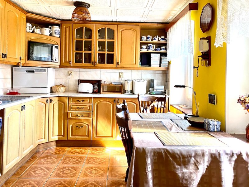 kuchyňská linka se spotřebiči, dřevěný stůl se židlemi, žluté zdi, na stěně jsou hodiny