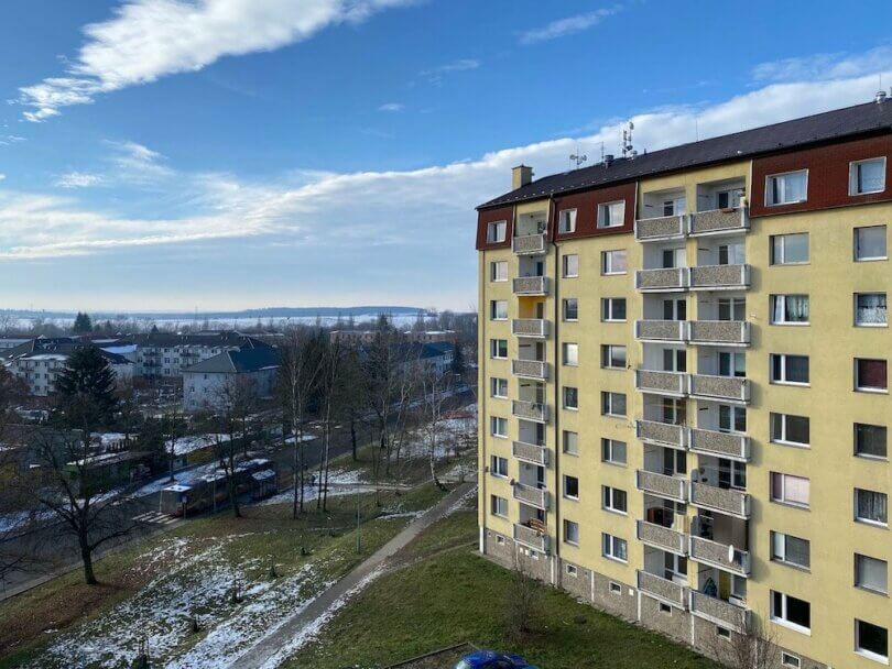 zateplený panelový dům s lodžiemi, silnice, chodník, zbytky sněhu a modrá obloha